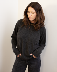 A woman with long brown hair stands against a white wall, looking down with her hands in her dark pants pockets. She wears the Autumn Cashmere Relaxed Fit Raglan Crew W/ Rib Detail in Pepper, appearing thoughtful and introspective.