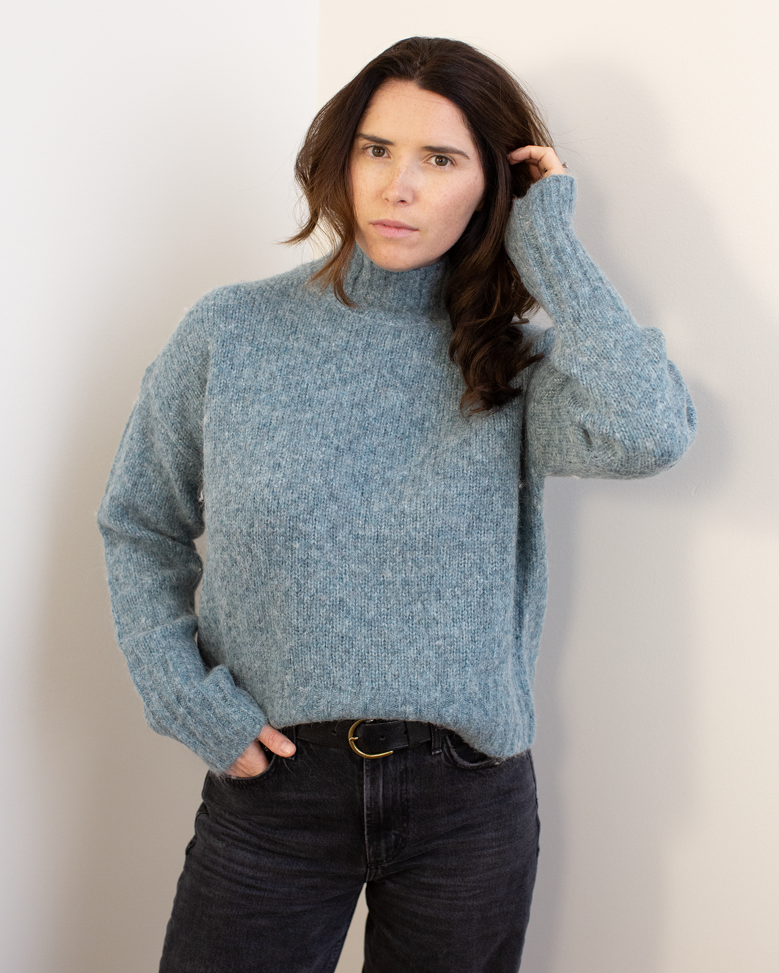 A person with long brown hair wears the Hartford Myac Turtleneck Sweater in Jade and black jeans, standing against a white wall with one hand in their pocket and the other touching their hair, looking directly at the camera.