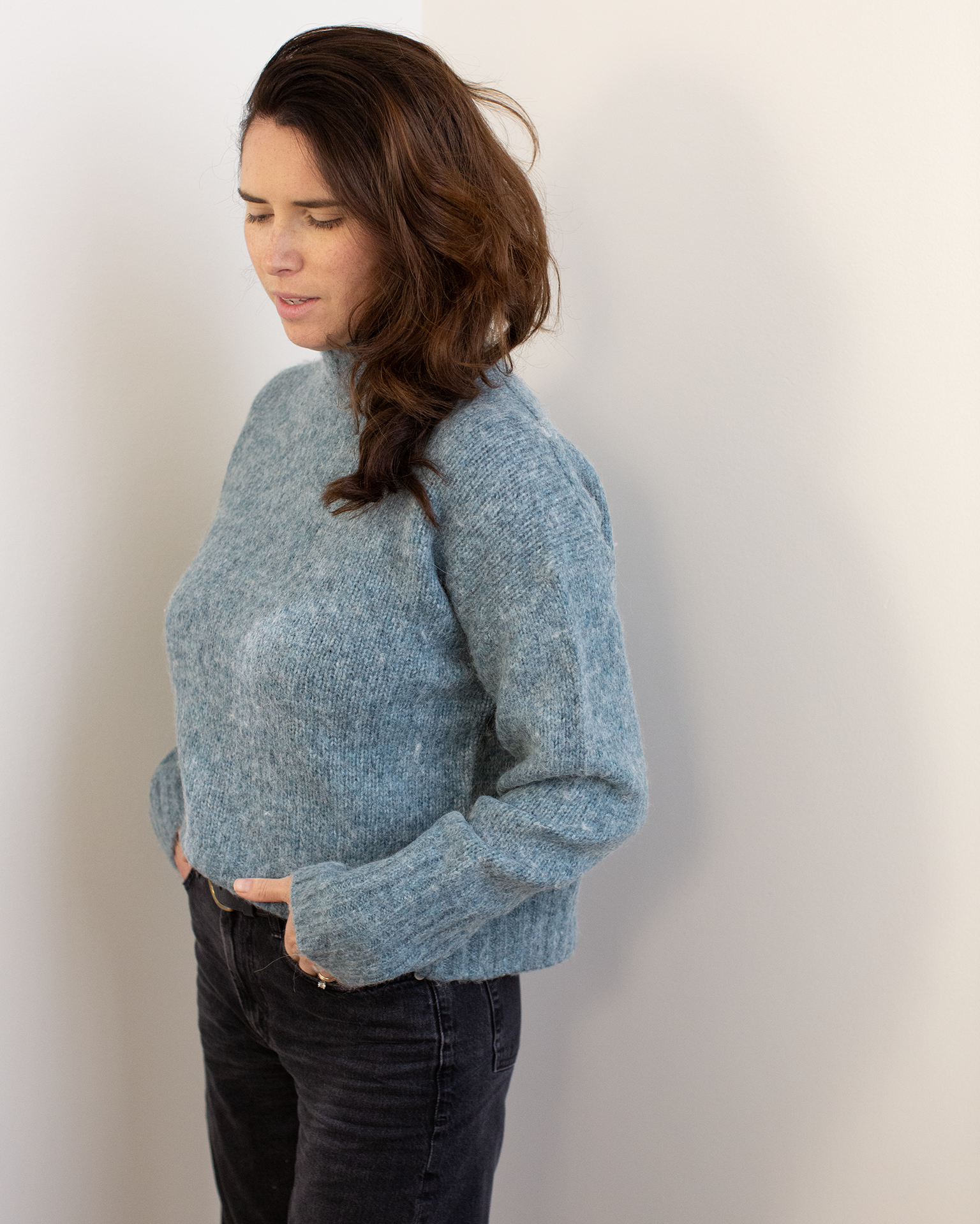 A woman with long brown hair stands against a white wall, wearing the Hartford Myac Turtleneck Sweater in Jade with black jeans, her hands in her pockets and looking slightly downward.