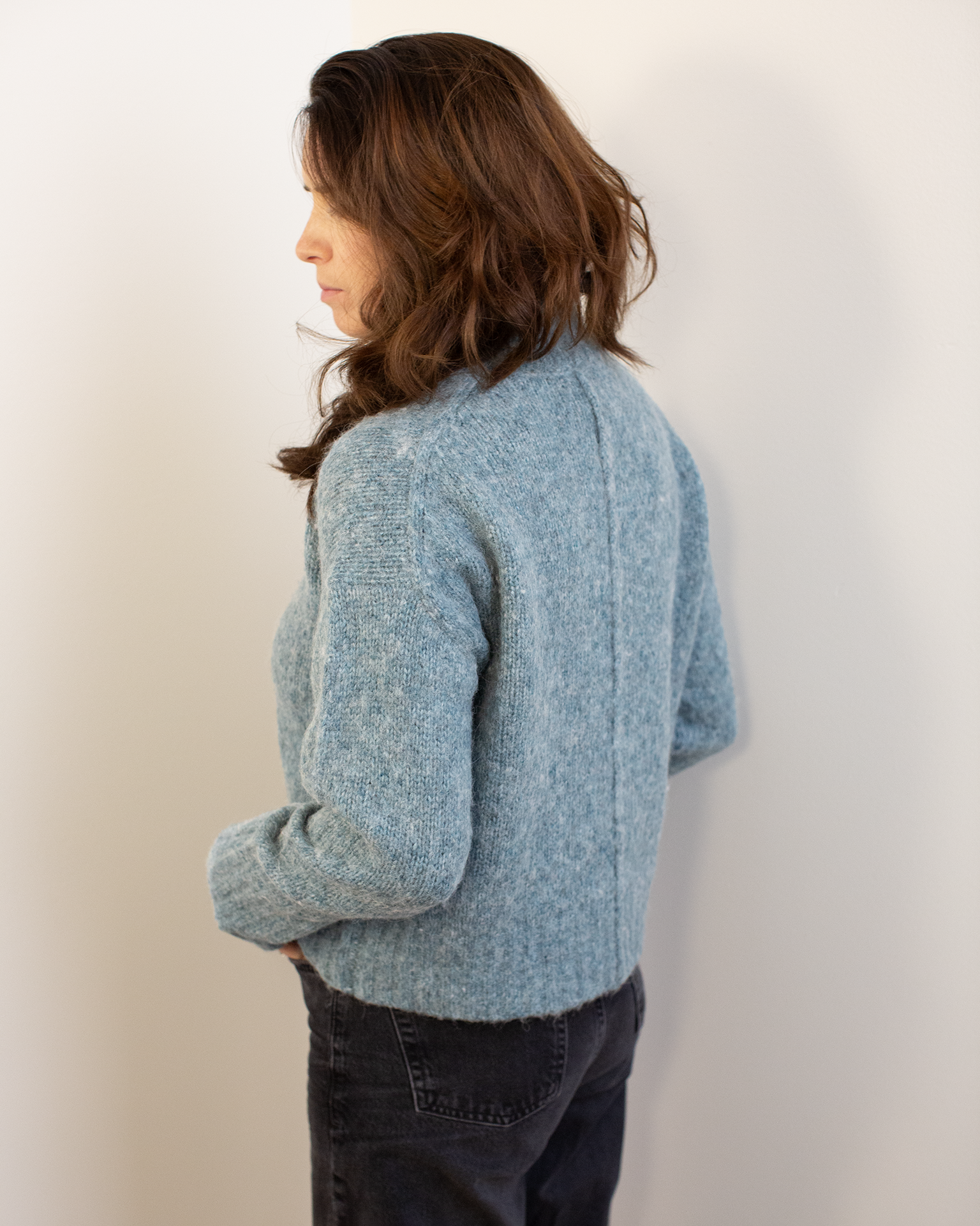 A woman stands by a wall, stylishly dressed in a Hartford Myac Turtleneck Sweater in Jade—a cozy wool-alpaca blend pullover that adds warmth and elegance to her look.