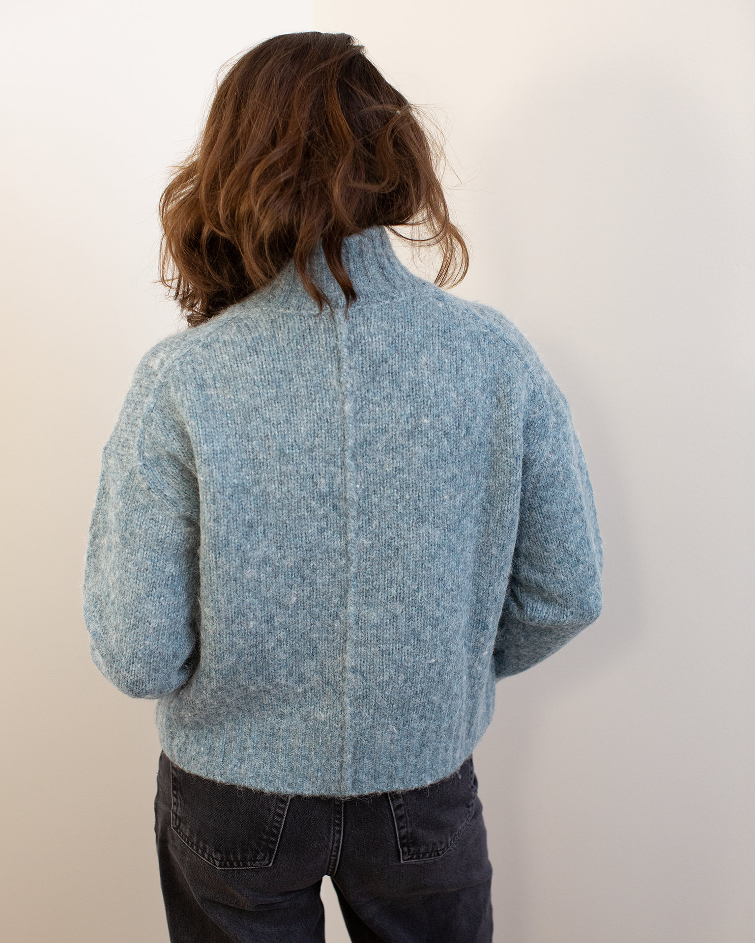A person with wavy brown hair, wearing the Hartford Myac Turtleneck Sweater in Jade and dark jeans, stands facing away from the camera against a plain white wall.