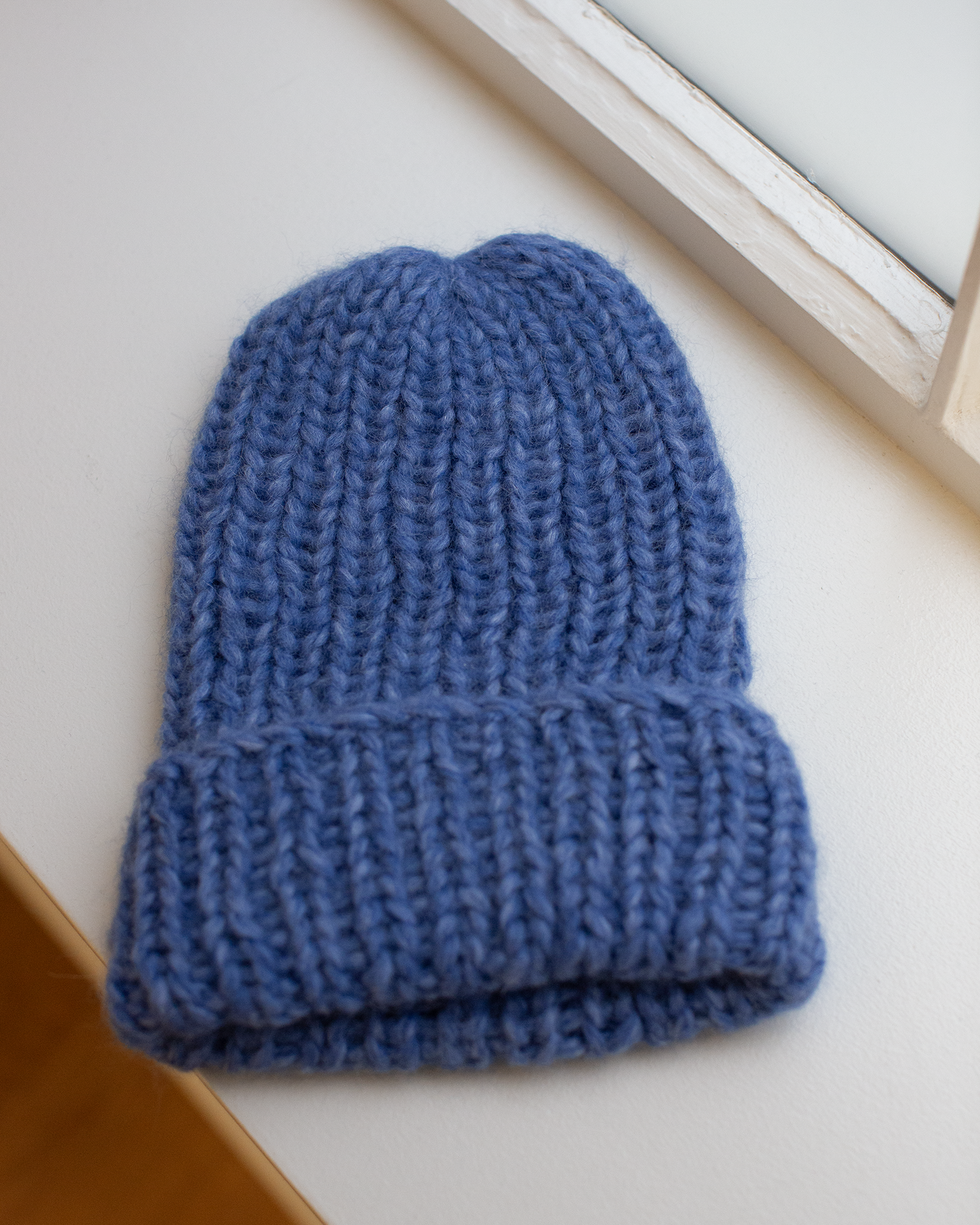 The Chunky Alpaca Hat in Blue Melange by Maiami, handknit from soft alpaca, rests on a white windowsill beside a matching white-framed window.
