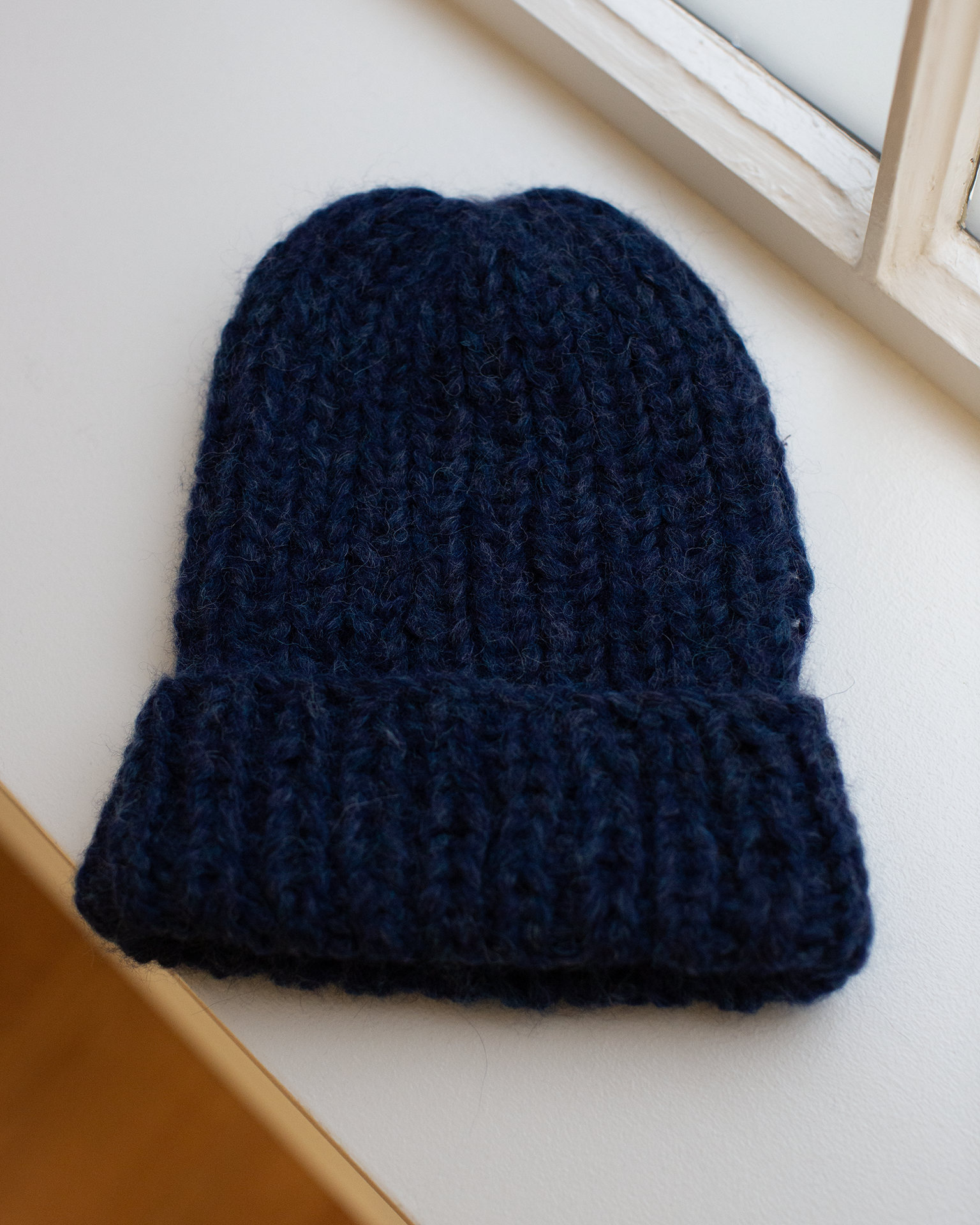 The Maiami Chunky Alpaca Hat in Denim, handknit and thick with a folded brim, rests on a white windowsill near a window.