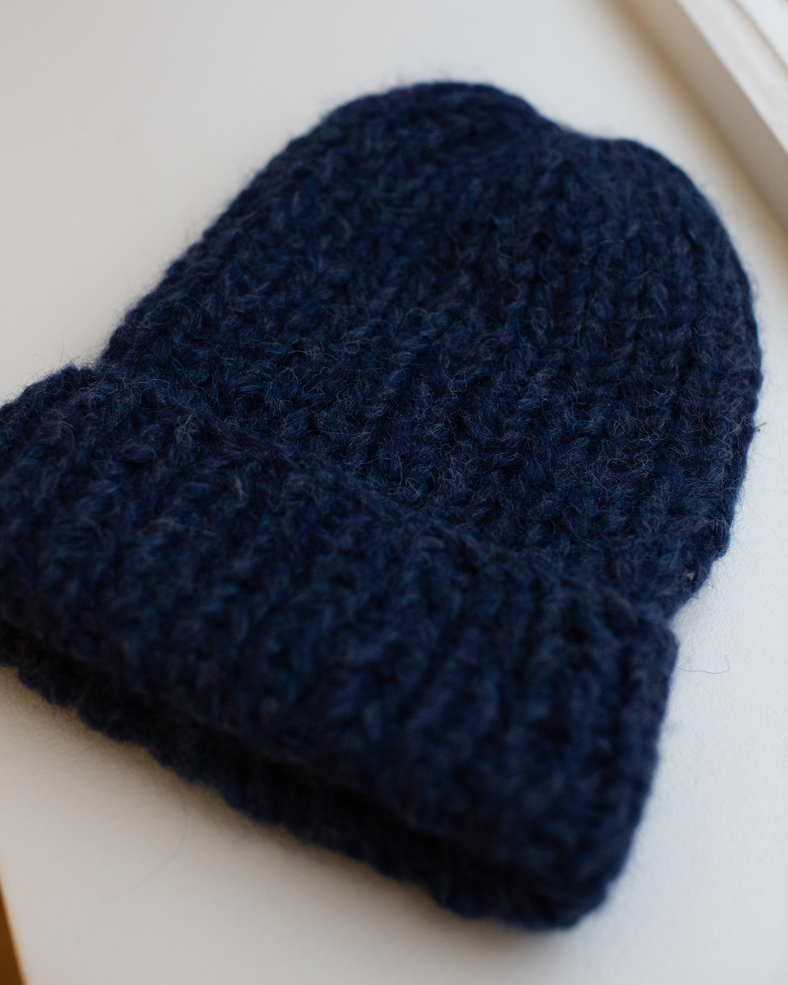 The Maiami Chunky Alpaca Hat in Denim, a dark blue handknit beanie with a thick alpaca texture and folded brim, is displayed flat on a light surface by the window.