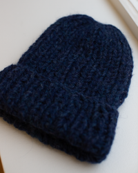 The Maiami Chunky Alpaca Hat in Denim, a dark blue handknit beanie with a thick alpaca texture and folded brim, is displayed flat on a light surface by the window.
