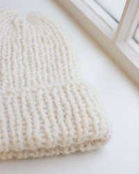A close-up of Maiami's Chunky Alpaca Hat in Creme, a handknit, off-white beanie, rests on a white surface by a sunlit window.
