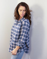 A woman with wavy brown hair wears the Frank & Eileen Eileen Relaxed Button Up Shirt in Grey & Red Plaid and light blue jeans, standing against a white wall and looking down to her left with a calm expression.