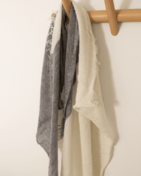 The Grisal Love Duo Mini scarf in Milk and Heather Grey, featuring color blocks and frayed edges, hangs on a light wooden hook against a white wall.