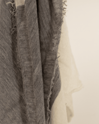 Close-up of overlapping pieces of frayed grey and off-white fabric, resembling the Grisal Love Duo Mini in Milk X Heather Grey scarf, with textured edges hanging against a neutral backdrop.