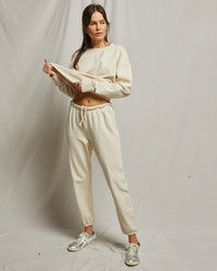 A woman stands against a neutral backdrop wearing perfectwhitetee's cream Johnny French Terry Easy Sweatpant in Sugar and a matching sweatshirt, lifting it to reveal her midriff and looking to the side, paired with silver sneakers.
