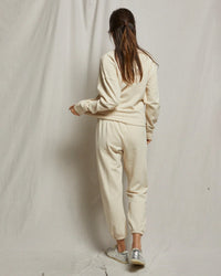 A woman with long brown hair, wearing perfectwhitetee's Johnny French Terry Easy Sweatpant in Sugar, stands with her back to the camera against a plain light backdrop, paired with silver sneakers.