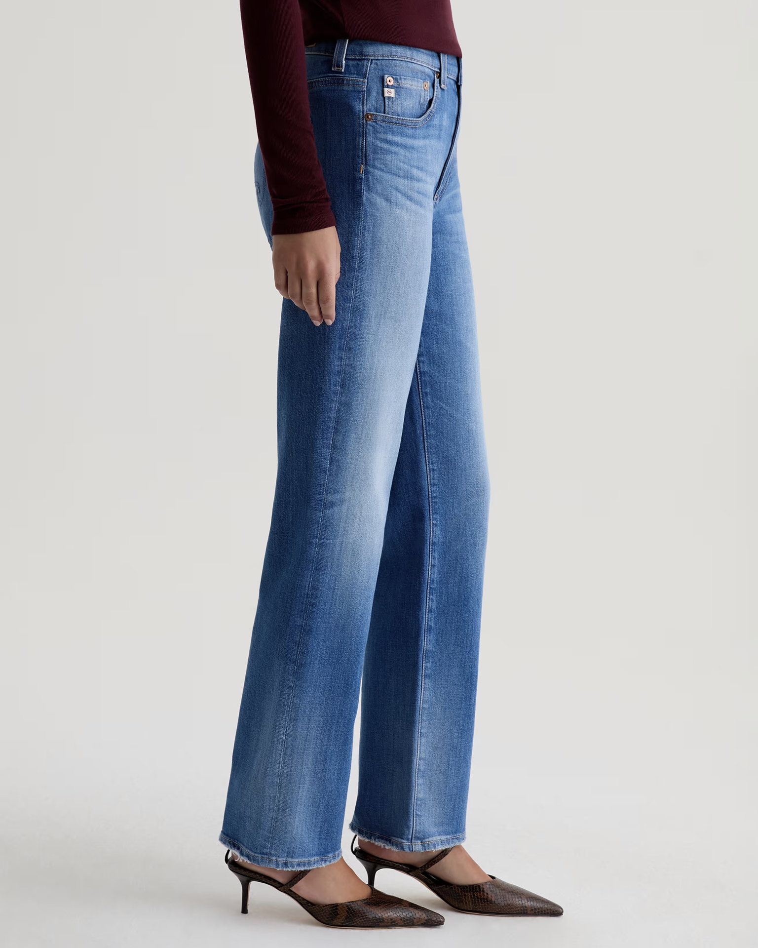A person stands against a plain light background wearing AG Jeans' The Brinley Straight in 13Ys Allure, a long-sleeve burgundy top, and brown pointed-toe heels.