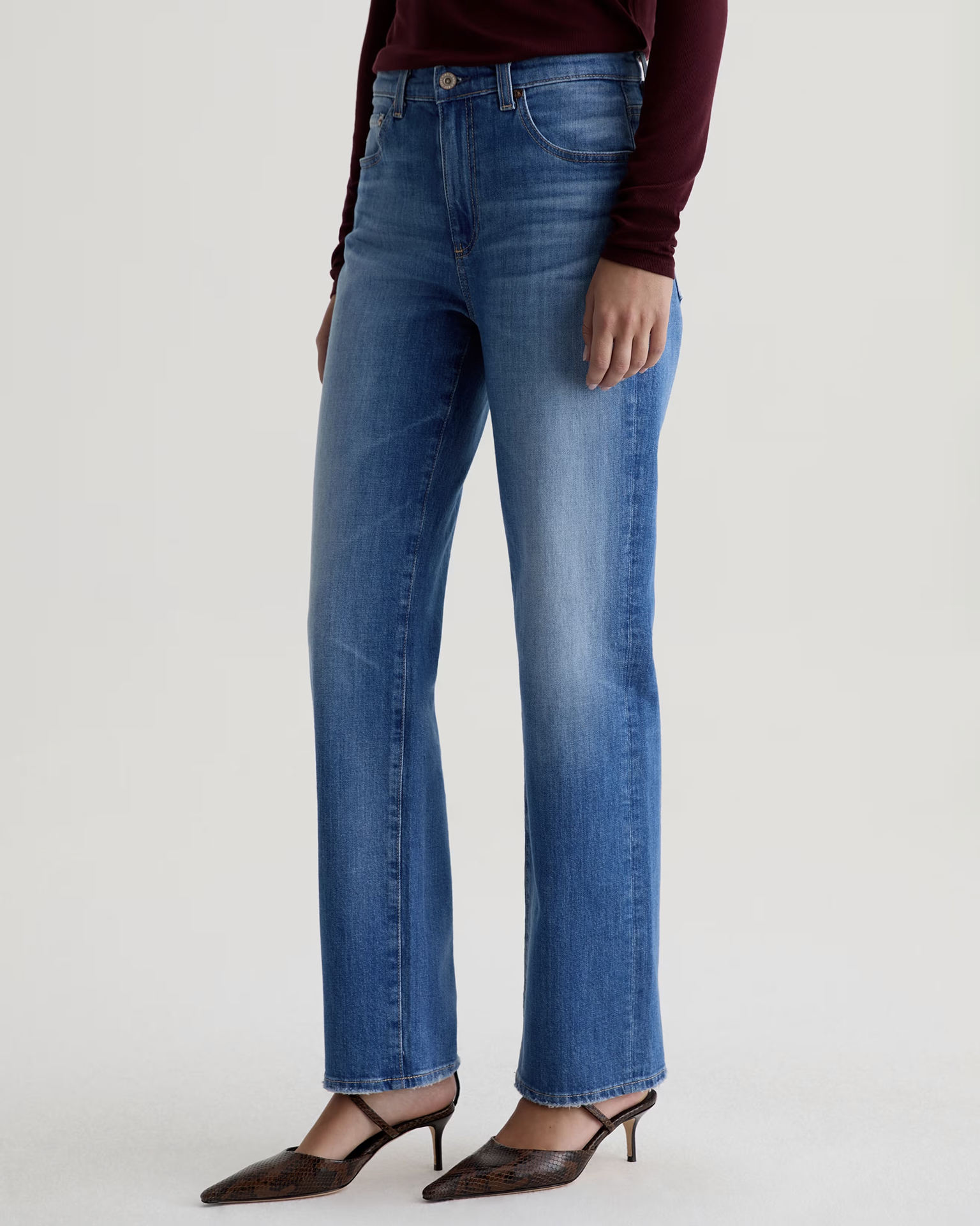 A person wearing The Brinley Straight in 13Ys Allure by AG Jeans, a long-sleeve burgundy top, and pointed-toe heels stands against a plain light background. The image is cropped from the shoulders down.