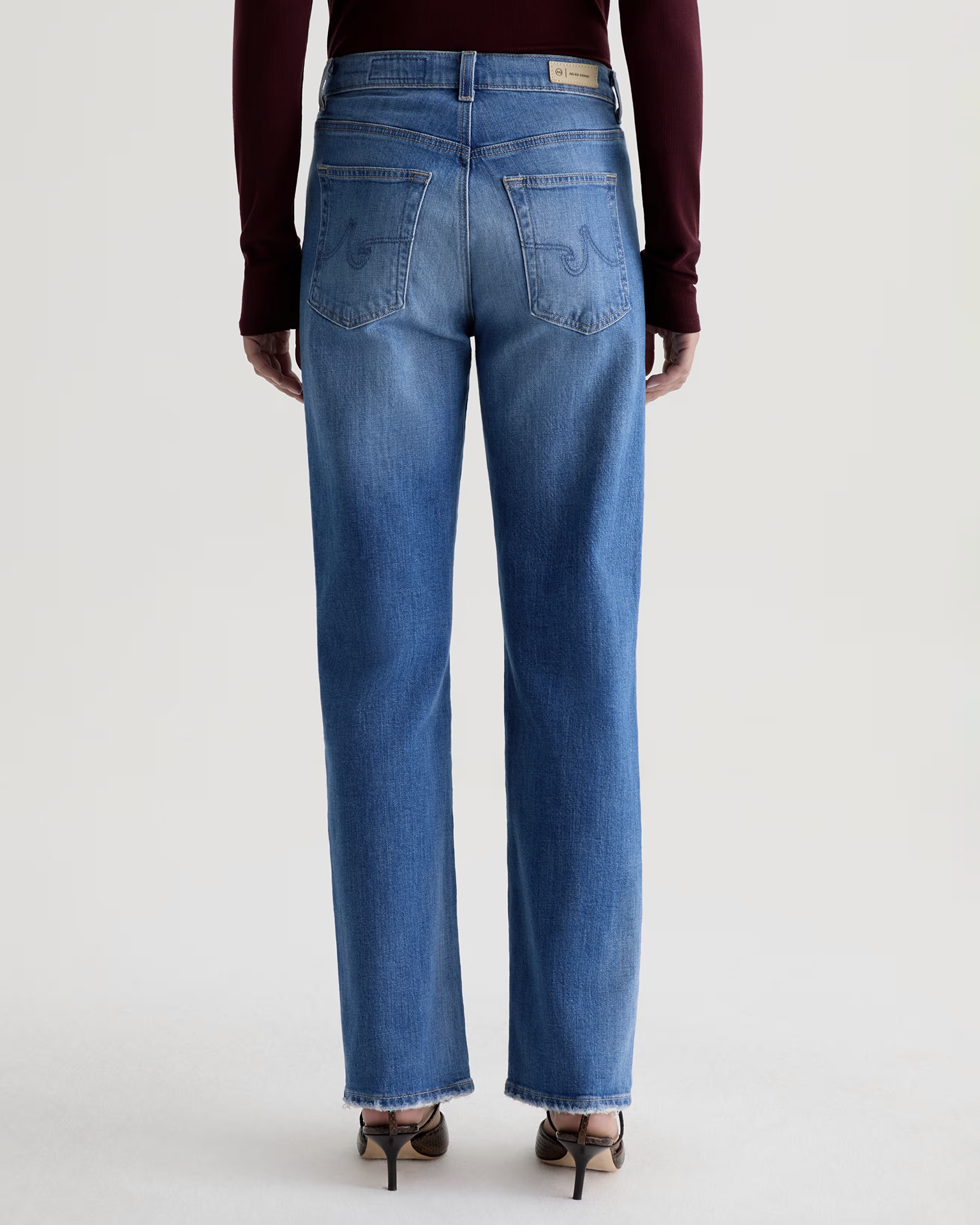 A person is shown from the back wearing AG Jeans' The Brinley Straight in 13Ys Allure, styled with a maroon long-sleeve top and heeled sandals against a plain light background.