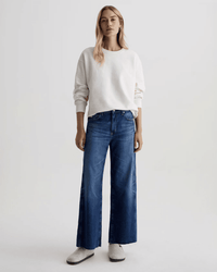 A woman stands facing forward against a plain light background, wearing a white sweatshirt, AG Jeans Saige Wide Leg Crop in Watermist, and light gray slip-on shoes.
