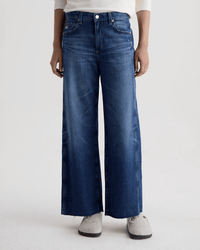 Model wears AG Jeans' Saige Wide Leg Crop in Watermist, a medium blue jean with a raw-cut hem, styled with a white top and grey slip-ons. Only their lower half is visible against a plain light background.