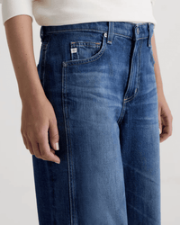 A person wears AG Jeans Saige Wide Leg Crop in Watermist, medium blue with a raw-cut hem and a white long-sleeve top, shown waist to mid-thigh, one hand resting by their side.