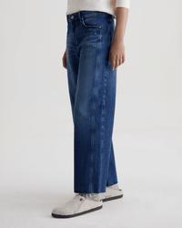 A person wearing a white long-sleeve shirt, AG Jeans Saige Wide Leg Crop in Watermist, and gray slip-on shoes stands against a plain light background.