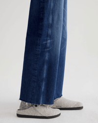 A person is wearing AG Jeans’ Saige Wide Leg Crop in Watermist, featuring a raw-cut hem, paired with light gray felt slippers with black soles, and standing on a light-colored surface.