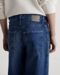 A person shown from the back wears AG Jeans' Saige Wide Leg Crop in Watermist medium blue, paired with a cream long-sleeve top. The jeans have large back pockets, a leather waistband patch, visible stitching, and a raw-cut hem.