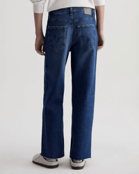 A person stands with their back to the camera, wearing AG Jeans' Saige Wide Leg Crop in Watermist, featuring a medium blue wash, raw-cut hem, back pockets, a white top, and gray slippers against a plain light background.