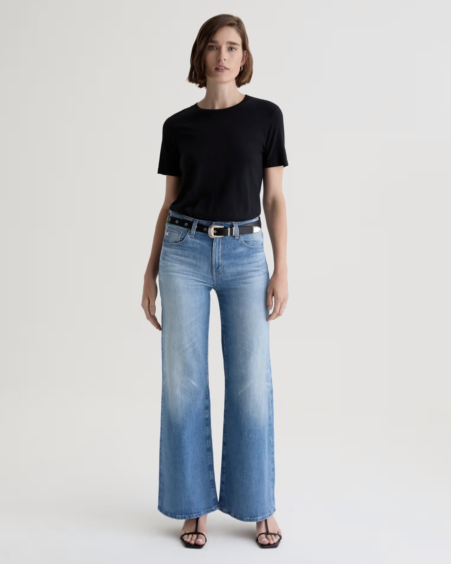 A person stands facing forward, wearing a black short-sleeve t-shirt, the AG Jeans Saige Wide Leg in 18Ys Bisou with a black belt, and black heeled sandals. The background is plain and light-colored.