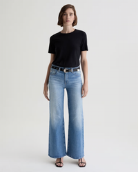 A person stands facing forward, wearing a black short-sleeve t-shirt, the AG Jeans Saige Wide Leg in 18Ys Bisou with a black belt, and black heeled sandals. The background is plain and light-colored.