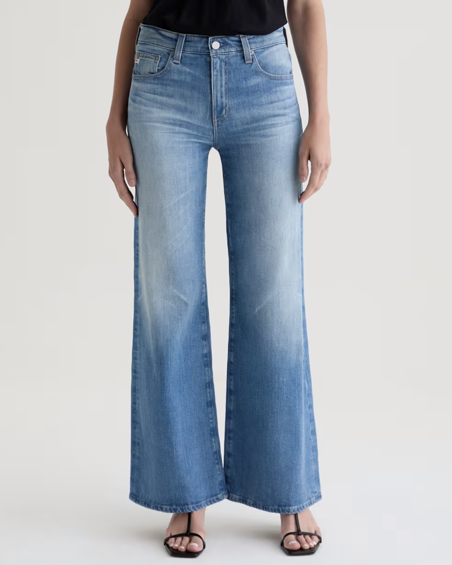 A person wears AG Jeans' Saige Wide Leg in 18Ys Bisou—light blue, high-rise, wide-leg jeans with subtle fading—paired with a black top and black heeled sandals, standing against a plain background.