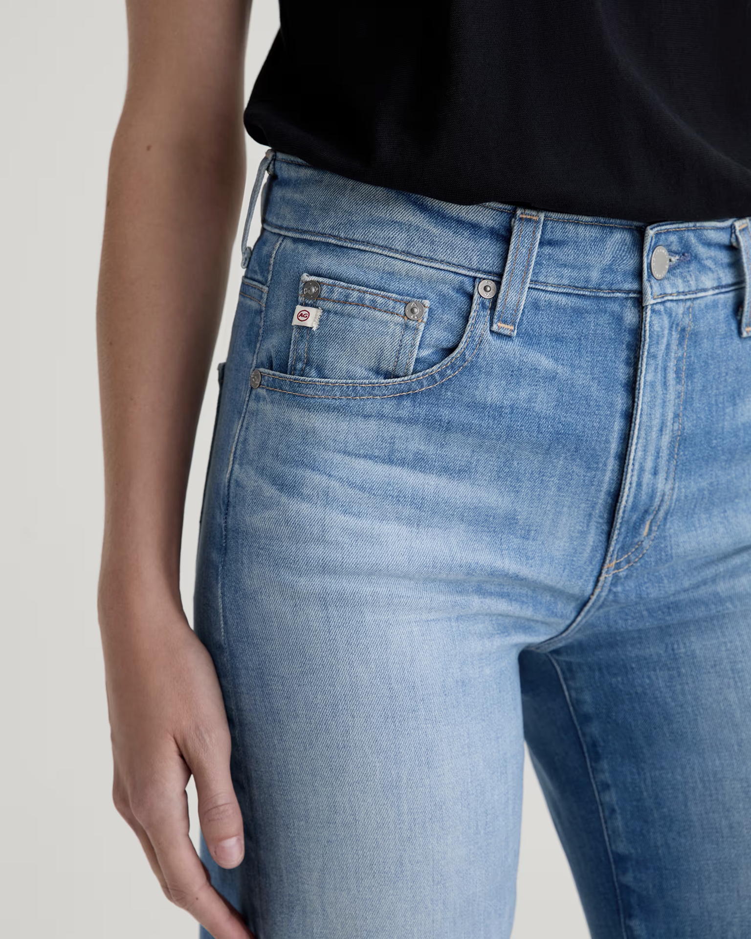 A person wears AG Jeans' Saige Wide Leg in 18Ys Bisou—light blue, high-rise jeans—paired with a black top. Their left arm is relaxed by their side, showcasing the jeans’ fit and unique fading details.