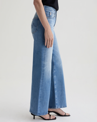 A person wearing AG Jeans Saige Wide Leg in 18Ys Bisou, a black sleeveless top, and black heeled sandals stands against a plain light background, shown only from the waist down.