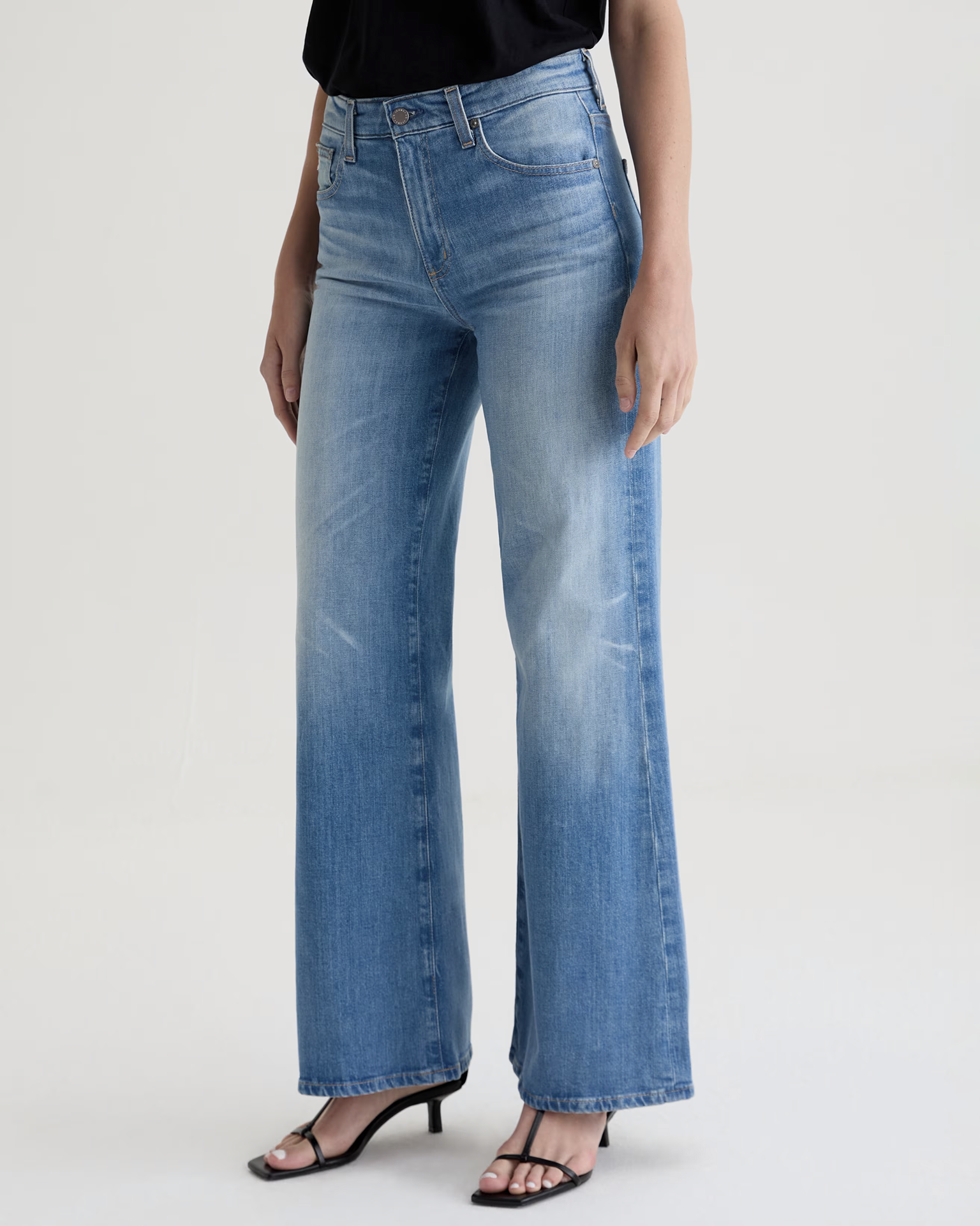 A person wears AG Jeans' Saige Wide Leg in 18Ys Bisou and black open-toe heeled sandals, standing against a plain light background with only their lower body visible.