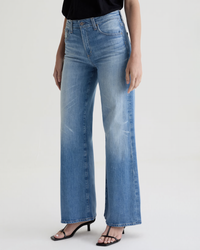 A person wears AG Jeans' Saige Wide Leg in 18Ys Bisou and black open-toe heeled sandals, standing against a plain light background with only their lower body visible.