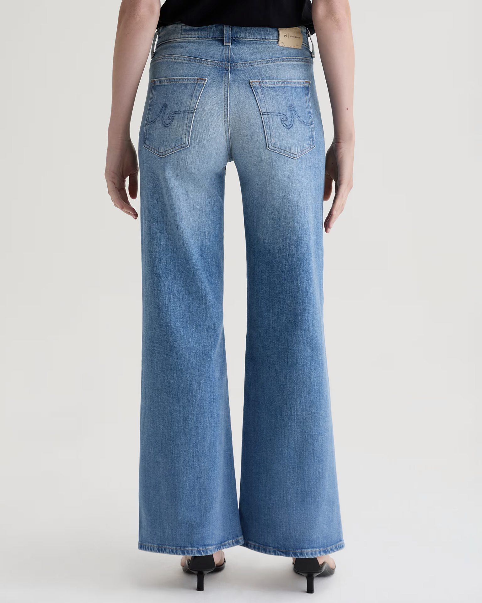A person wearing AG Jeans' Saige Wide Leg in 18Ys Bisou—a light blue, high-rise, wide-leg cropped jean—paired with black heeled sandals, shown from the back against a plain light background.
