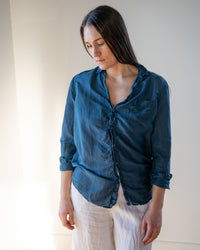 A woman with long brown hair stands indoors, looking down with a neutral expression. She wears the LLEA BLUE Carrie Blouse in Moonlit Ocean cotton silk and light-colored pants, illuminated by natural light from the left.