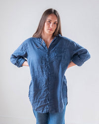 A woman with long brown hair wears the LLEA BLUE Ciel Tunic Blouse in Moonlit Ocean Linen and jeans, standing with hands on her back and a neutral expression against a plain light background.