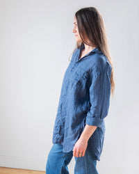 A woman with long brown hair stands in profile against a light wall, wearing the LLEA BLUE Ciel Tunic Blouse in Moonlit Ocean Linen styled as a blue button down shirt, paired with blue jeans.