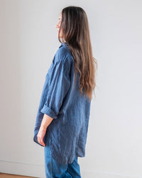 A person with long brown hair stands in profile, facing left, wearing the LLEA BLUE Ciel Tunic Blouse in Moonlit Ocean Linen and blue jeans against a plain light-colored wall.