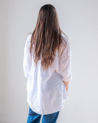 A person with long brown hair, wearing the LLEA BLUE Ciel Tunic Blouse in White Linen and blue jeans, stands facing away from the camera against a plain light-colored wall.