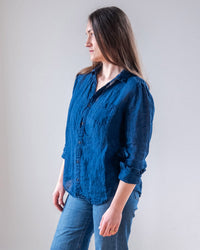 A woman with long brown hair stands sideways, wearing the LLEA BLUE Carrie Fitted Blouse in Indigo and blue jeans. She showcases a stylish yet relaxed look against a plain light background.