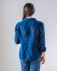 A person with long brown hair, wearing the LLEA BLUE Carrie Fitted Blouse in Indigo and slim blue jeans, stands facing away from the camera against a plain light background.