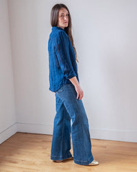 A woman with long brown hair stands in a minimalist room, wearing the LLEA BLUE Carrie Fitted Blouse in Indigo and wide-leg jeans. She looks over her shoulder at the camera, one foot forward to accentuate her slim figure.