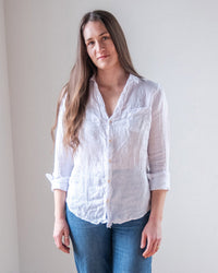 A woman with long brown hair wears the LLEA BLUE Carrie Fitted Blouse in White Linen and blue jeans, standing against a plain light wall and looking at the camera with a slight smile.