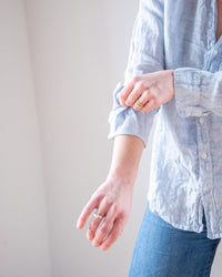 A person in the LLEA BLUE Carrie Fitted Blouse in Aqua Chambray Linen with jeans rolls up their sleeve, revealing ring-adorned hands against a plain light background.