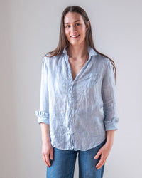 A woman with long brown hair, wearing the LLEA BLUE Carrie Fitted Blouse in Aqua Chambray Linen and blue jeans, stands against a light background, smiling at the camera.