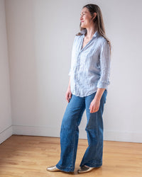 A woman stands indoors on a wooden floor, smiling to the side and wearing the LLEA BLUE Carrie Fitted Blouse in Aqua Chambray Linen with blue wide-leg jeans and gold shoes.