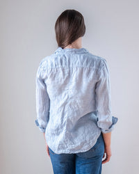 A person stands with their back to the camera, wearing the LLEA BLUE Carrie Fitted Blouse in Aqua Chambray Linen and blue jeans. Their long brown hair is pulled to one side against a plain, light background.