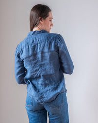 A person with long brown hair, seen from behind against a plain light background, is wearing the LLEA BLUE Carrie Fitted Blouse in Moonlit Ocean Linen and blue jeans.