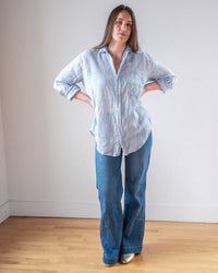 A woman stands indoors on a wooden floor, wearing the LLEA BLUE Alix Top in Aqua Chambray Linen and wide-leg blue jeans, hands on hips, with long hair down, against a plain white wall.