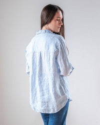 A woman with long brown hair, wearing the LLEA BLUE Alix Top in Aqua Chambray Linen and blue jeans, stands facing away from the camera and looks over her left shoulder against a plain light background.