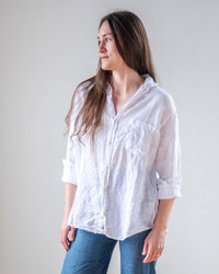 A person with long brown hair, wearing the LLEA BLUE Alix Top in White Linen and blue jeans, stands against a plain light background, looking slightly to the side and smiling gently.